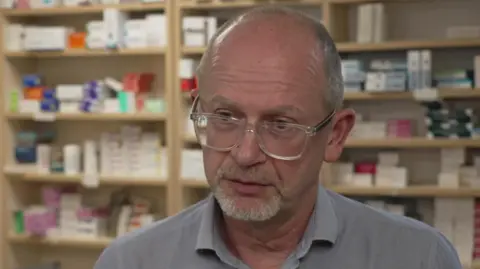 A man wearing clear rimmed rectangle glasses and a light blue collared shirt standing in a pharmacy with shelves of drugs behind him.