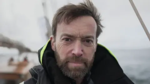 Turn to Starboard Paul Miller smiling at the camera. He has a moustache and beard and is stood on a ship.