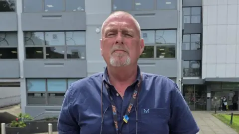 Geoff is standing outside an office building wearing a blue shirt and looking at the camera.