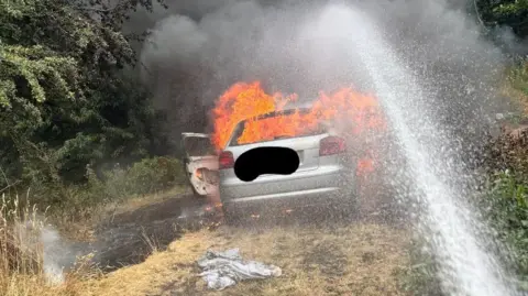 Shropshire Fire & Rescue Service A silver car engulfed in flames on the grass.