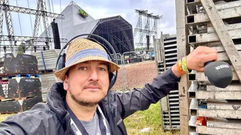 Selfie of Andy Bennett. He has a sun hat and headphones on and he is holding a BBC microphone. Andy is stood in front of the Pyramid Stage on site at Glastonbury Festival. 