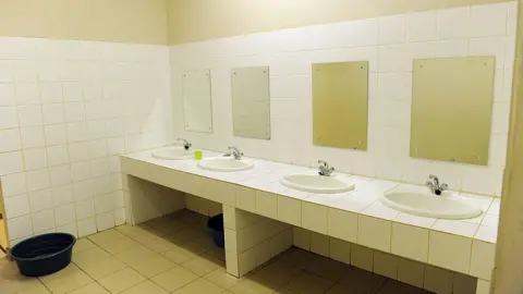 Getty Images A tiled bathroom with four sinks and the same amount of rectangular mirrors fixed to the wall above. A plastic bucket is placed on the floor a few feet away from sinks. 