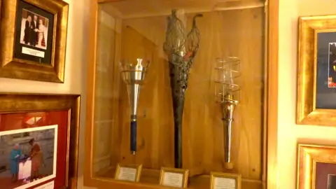 Qays Najm/BBC Ceremonial torches in an enclosed glass and wooden-framed case are mounted on a wall. Adjacent are gold-framed photos including one of the late Queen Elizabeth II with a Beefeater in his red and gold outfit.