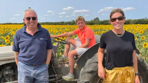 A man and woman stand in front of a vintage tractor, parked in a field of sunflowers. A young boy sits on the tractor seat.