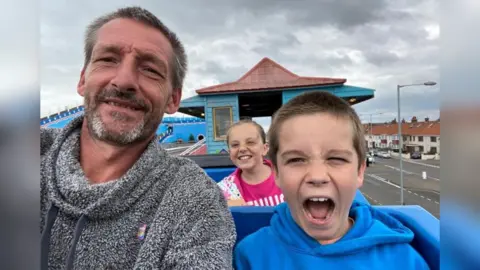 The Coleman Family Jason Coleman, Freddie Coleman and a young girl sat on what appears to be a roller coaster at a theme park. Jason is wearing a gray and white hoodie, has short gray hair, and is smiling. Freddie is under 15 here. He has short brown hair, is wearing a blue hoodie, and has his mouth open in excitement. The girl smiles while wearing a pink t-shirt. She has light hair that has partially fallen into her face.