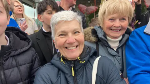 Standing in front of a number of other people, a woman smiles a broad smile. She has short silver hair and is wearing a blue coat. The white strap of a bag is over her shoulder, her arms are folded.