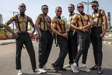 Olympia De Maismont / AFP A group of men participating in the Calabar Carnival pose for a photo wearing black pants, black belts and sunglasses. Their chests are bare and covered with a golden glow. Some wear black ties.