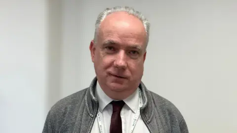 Kate Moser Andon/BBC Angus Ellis headshot, he has a neutral expression, against a white wall background.