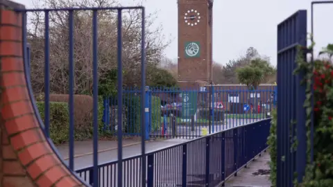 View through blue metal railings towards a the Fobbing Academy's school site, with a brick clock tower and Mossbourne signage visible in the distance.