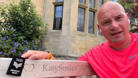 Matthew Miles Image of a man wearing a red t-shirt and yellow watch. He is sitting on a bench which has two carved hearts and the words 'Katy Smiles'. The man is holding a police sergeants badge. In the background can be seen a stone building with leaded light windows and shrubbery in front.