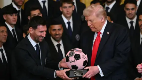 Lionel Messi handing Donald Trump a pink and black soccer ball trophy