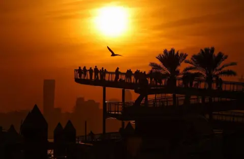 A group of people gather on a walkway raised in Dubai Creek Harbor at the Fadel Army/AFP sunset. A big bird is flying over them. The distance has skyscraper buildings and the sunset orange puts everything in the silhouette.