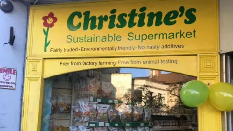 Chris Giles The front of Chris' shop, with large green letters reading "Christine's sustainable supermarket". The shopfront is bright yellow around a large window and there are two balloons tied to the right of it.
