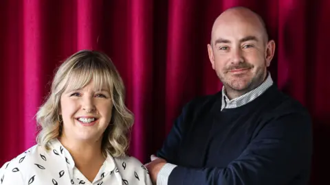 BBC Tara Mills and Declan Harvey standing in front of a red curtain. Tara is wearing a white blouse and Declan has a black jumper on. Both are smiling towards the camera.