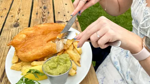 A woman eats a plate of fish and chips with some mushy peas in a pot next to her - her plate resting on a wooden table in an outdoor restaurant.