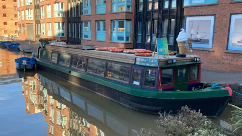 The outside of the narrowboat, which is green and next to a number of flats along the waterside. There are a number of life rings on the roof and large model of an ice cream. 