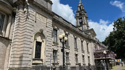 The large building of Torquay Town Hall which has decorative window details and railings in front. The pale, stone coloured building has a clock tower showing the time 12:00.