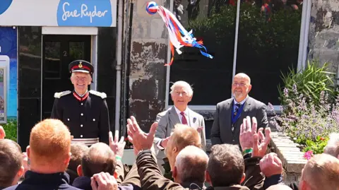King Charles throws a bright coloured ball with ribbons into a crowd of men waiting to grab it