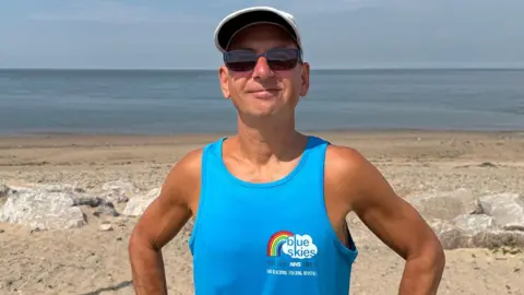 Peter Franklin stands on a beach in the sunshine with his hands on his hips. He is wearing a blue vest top, a white baseball hat and sunglasses and is smiling