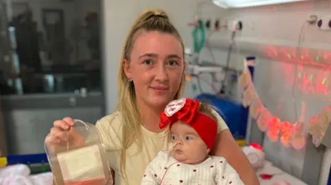 Family photo Dolcie-Mae's mum holds a blood bag and she is wearing a yellow top. Dolcie-Mae is being held in her arms with wispy brown hair, a red bow on her head and a white baby grow with love hearts on it.