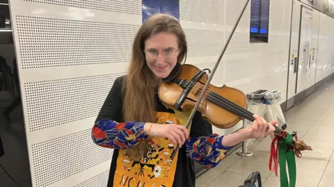 Natalie Russo, a woman with long hair and a colourful top holding a violin decorated with red and green bows