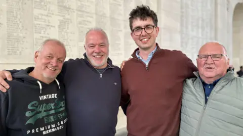 Four men are smiling at the camera. They are standing in front of a wall with names inscribed. They are all smiling and have their arms around each other. 