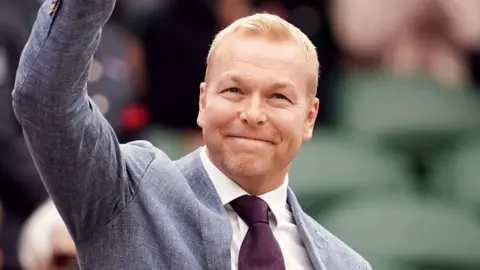 Pa media Sir Chris Hoy dalam setelan abu -abu dan Maroon Tie Smiles and Waves ke kamera