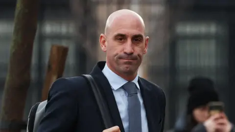 Reuters Luis Rubiales hold the strap of the backpack his is wearing over his right shoulder. He is wearing a suit and tie and is outside, on his way to the court in February 2025. In the background is a blurry woman wearing a beanie hat, holding her mobile phone up to her face to take a picture.