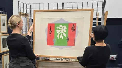 Two women are holding a large framed artwork in front of a metal display grid. The artwork features a bold abstract design with a central green panel flanked by red sections on each side. A white organic shape resembling a leaf or coral is in the centre, surrounded by smaller black leaf-like motifs. The frame is wooden, and additional framed pieces are visible behind and below the main artwork.