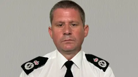 Chief Constable Ivan Balhatchet has cropped hair and serious expression. He is wearing a white short-sleeved police shirt with a black tie