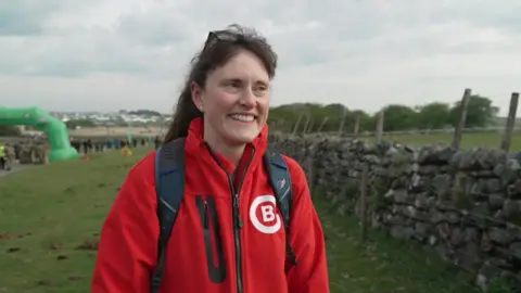 A woman in a red jacket and a black backpack is smiling. She is standing on the moors.