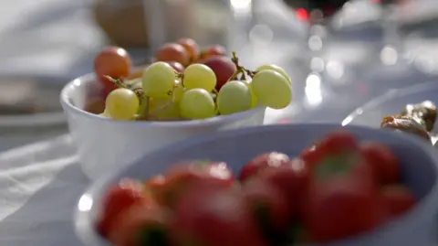 White wine glasses filled with green and red wine on a table covered with a white cloth. At long last, the other bowl contained a red strawberry, slightly out of focus. Latarsa shows blurred standware and other picnic items, showing outdoor dining settings in bright natural light.