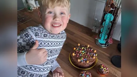 Alfie in a black and white top smiling and doing a thumbs up sign. He is smiling broadly lying on a brown floor beside chocolate cake decorated with colourful chocolate buttons. 