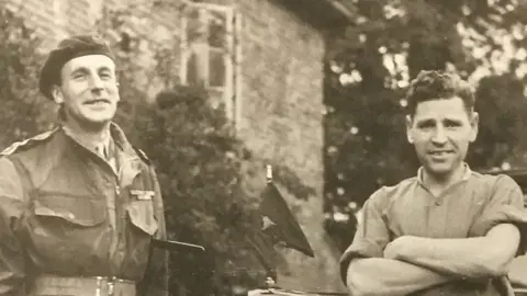 John Lloyd A black and white photo from spring 1945 in Germany. It shows Maj Gen Eric Bols on the left, who is wearing a beret and is in uniform, with his head back slightly as he smiles. On the right is Jack Lloyd, who is shorter, is looking directly at the camera with short hair and smiling slightly, longer at the top. He is wearing a collarless shirt with the sleeves rolled up and has his arms crossed. 