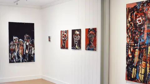 Linden Hall Studio Abstract paintings of people hung on gallery walls. Three small portraits with red or black backgrounds are hung next to each other depicting people from the shoulders up. A larger painting on the adjoining wall appears to show four or five white figures while another red, black and yellow painting is harder to discern.