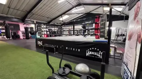 Charles Oliver A scene image of a boxing ring with gym equipment in the foreground