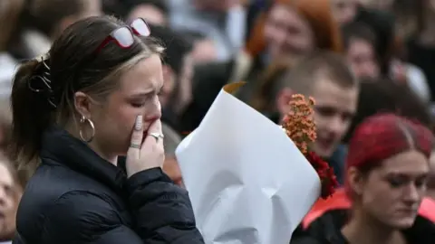 AFP A young woman wearing a black jacket holds a bouquet of flowers, one hand covering her mouth as she looks down, at a vigil with a crowd in the background