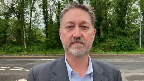 A man with grey hair and beard stares into the camera. He is wearing a grey jacket and light blue shirt. He's standing beside a road and there are trees in the background.