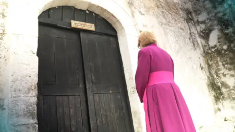 BBC News The Archbishop of Canterbury Dame Sarah Mullally, wearing purple robes, prays at the Door of No Return, a large black door, at Cape Coast Castle