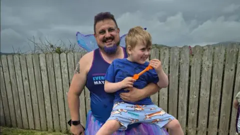 Lynda Davies Thomas Davies dressed in purple with fancy dress fairy wings, smiling, holding a smiling Lyidan.