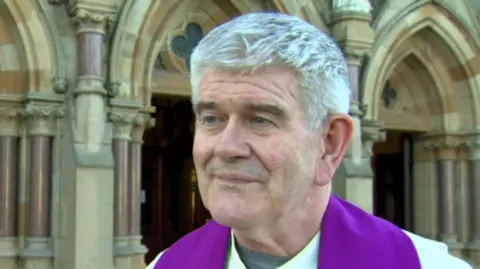 Fr. Ciarán O'Callaghan has abbreviated  achromatic  hair. He is looking to the near  of the surface  and is wearing a purple stole and achromatic  gown. 