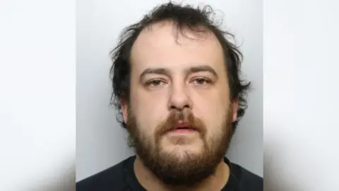 A police mugshot of a man with messy dark hair and a short beard. He is wearing a black T shirt.