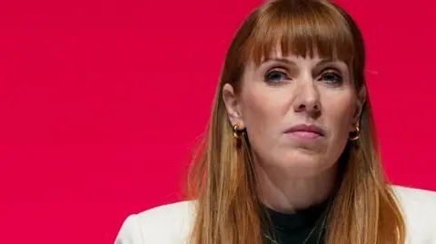 Getty Images Angela Rayner who has long, ginger hair and is wearing a white suit and gold hooped earrings, looks to her right in front of a red background
