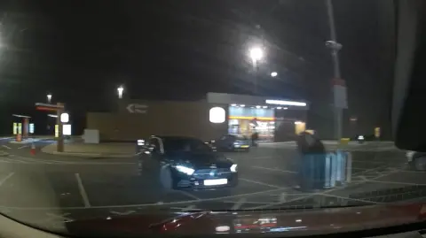 Jamie Andersen Dashcam image from a car at night driving into a Burger King Car park, with a black parked car in front of it and a full view of the restaurant, which has its lights on. The drive thru is to the left of the restaurant with a traffic cone outside.