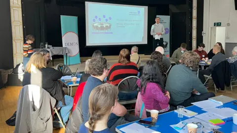 BBC/Richard Moss MP Lewis Atkinson stands on the stage of Seaton Delaval arts centre addressing a crowd of people sitting in the audience. He is holding a piece of paper and behind him is a slide projected on a screen with the title 'Your Voice on Migration'. The man has short grey hair and wears a light grey jumper over a white shirt and navy trousers.