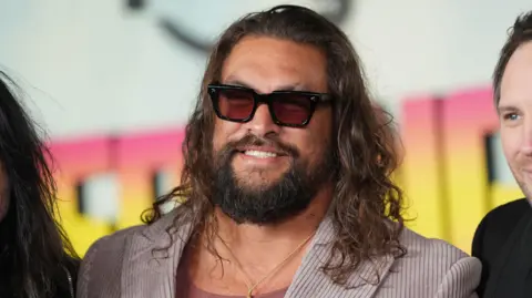 PA Media Momoa is stood in between two other people who are obscured in the image. He is wearing a pink corduroy suit and pink vest underneath and gold chain. He has his trademark long hair and beard and dark shades on.