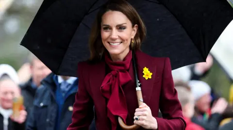 Catherine wearing burgundy with a yellow daffodil pinned to her blazer, holding a black umbrella, as people take pictures in the background