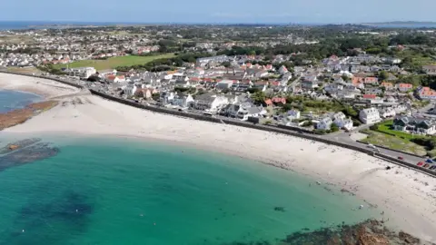 Houses and businesses are fronting onto a bay with a sandy beach and azure blue seas at Cobo in Guernsey.