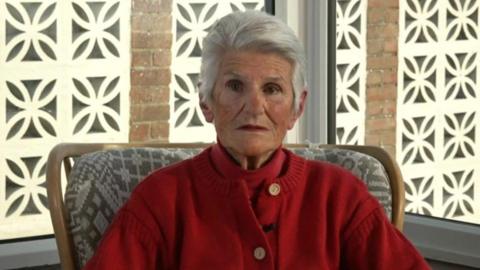 Veronica Smith, now an older woman, sits in a chair and wears a red jumper