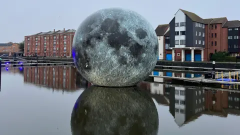 The Luke Jerram moon artwork is visible floating on the surface at Bridgwater Docks. Behind it various blocks of flats are visible and the water is calm